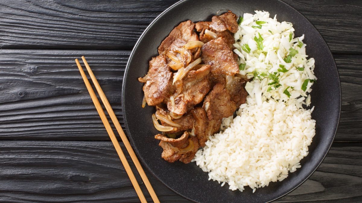 Gebratenes Schwein Shogayaki mit Reis und Krautsalat auf schwarzem Teller uns schwarzem Holztisch aus der Vogelperspektive. Links ein Paar Holzstäbchen.