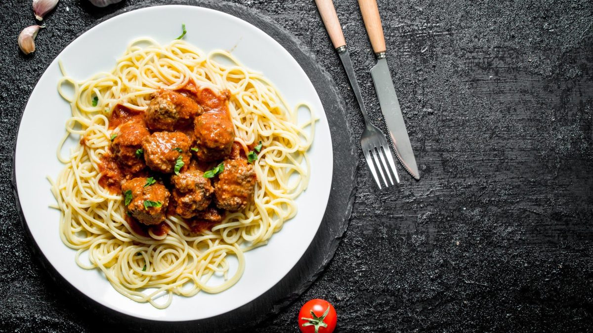 Spaghetti mit Fleischbällchen aus "Der Pate" auf weißem Teller und schwarzem Tisch aus der Vogelperspektive. Hackbällchen in Tomatensoße auf Spaghetti. Rechter Bildrand Messer und Gabel mit Holzgriff. Einzelne Tomate am unteren Bildrand zu sehen.