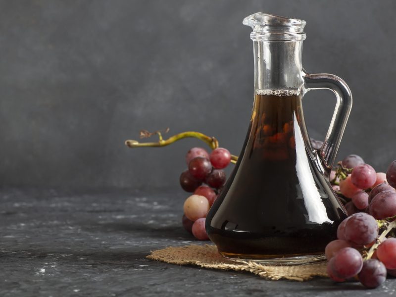 Eine Glaskaraffe mit einer dunklen Flüssigkeit steht auf dunklem Untergrund. Drumherum liegen Weintrauben.