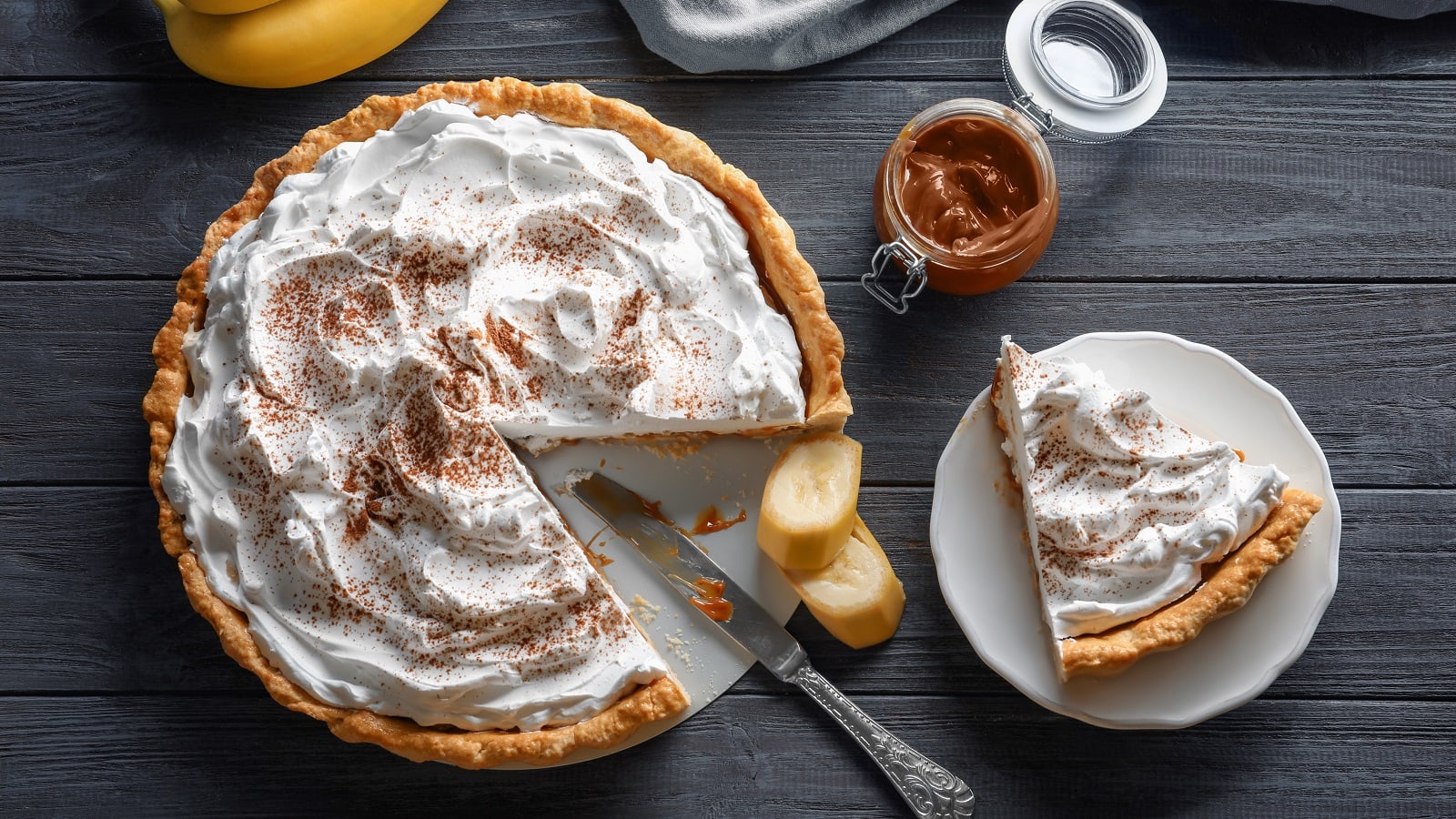 Ein angeschnittener Banoffee Pie wie aus dem Film Tatsächlich Liebe neben einem Messer, Bananenscheiben, einem Glas mit Karamell und einem Teller mit einem Stück des Kuchens vor einem grauen Hintergrund von oben.