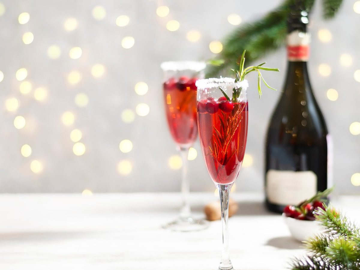 Zwei Gläser Cranberry Mimosas vor einem hellen Hintergrund, dahinter erkennt man unscharf eine Lichterkette und eine Champagnerflasche.