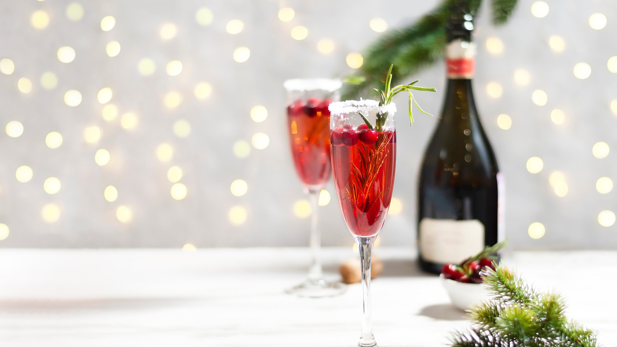Zwei Gläser Cranberry Mimosas vor einem hellen Hintergrund, dahinter erkennt man unscharf eine Lichterkette und eine Champagnerflasche.