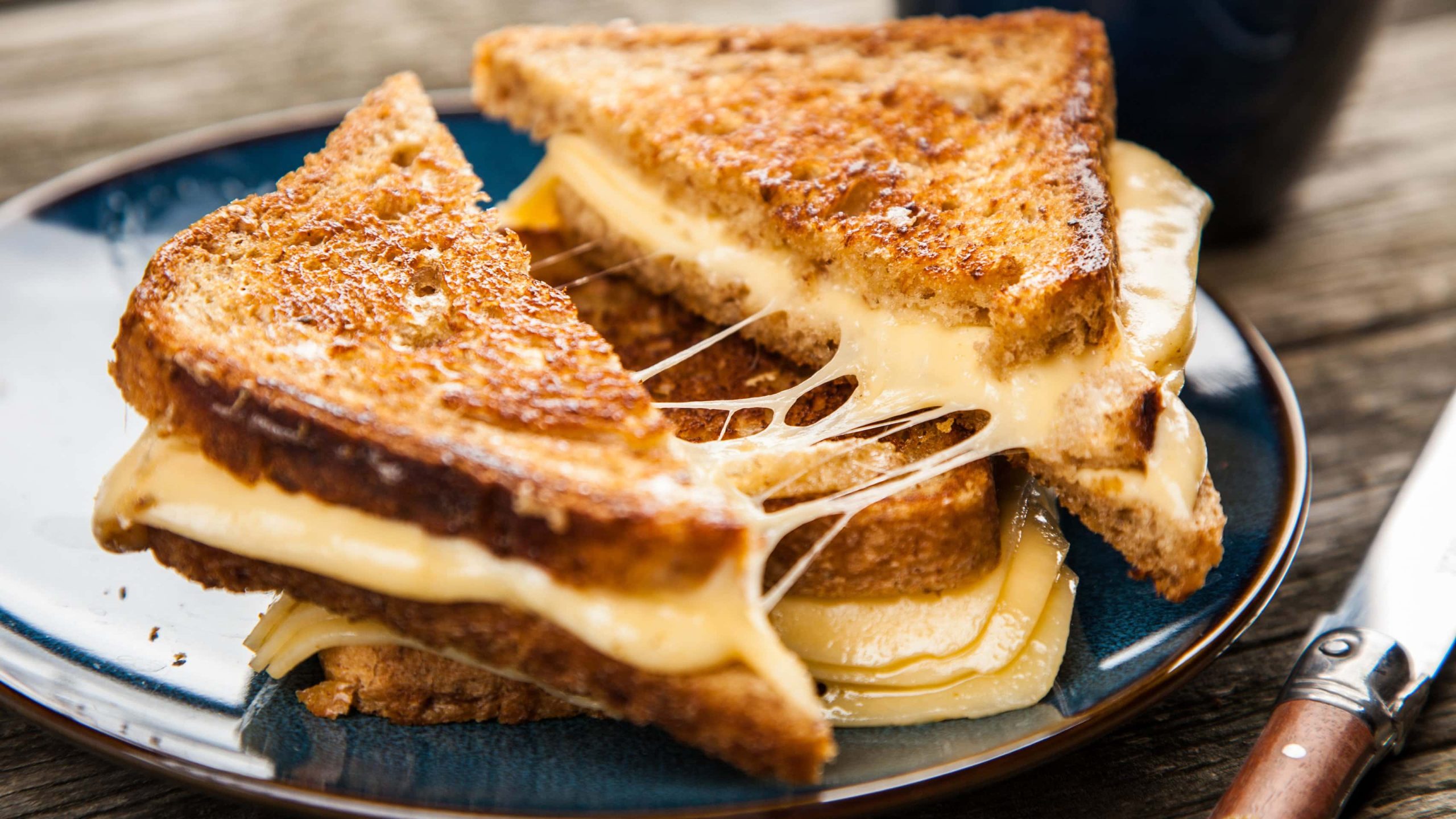 Zwei Grilled Cheese Sandwiches aus "Jane the Virgin" übereinander auf dunklem Teller auf Holztisch aus der Draufsicht. Das obere in der Mitte geteilt mit sich ziehendem geschmolzenem Käse. Rechts Messer mit Holzgriff.