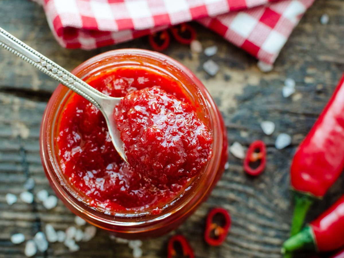 Draufsicht von oben: Ein Glas mit einer Tomaten-Chili-Marmelade steht auf einem Tisch. Daneben liegen zwei Chilischoten.. Die Marmelade heißt Red Hot Chili Jam..