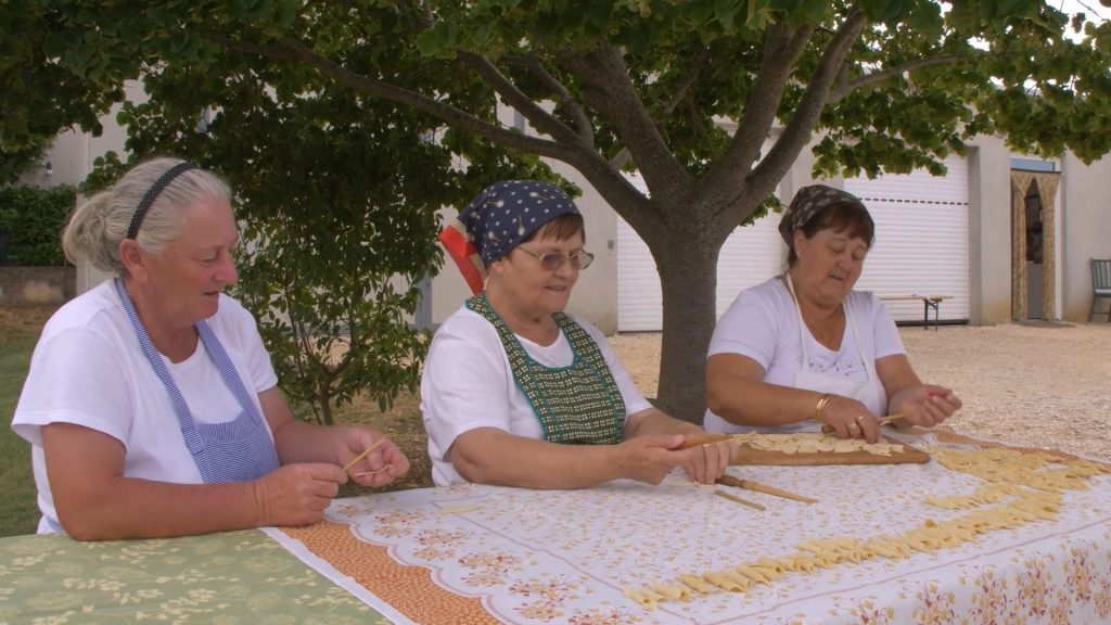 Drei Pasta Grannies sitzen draußen an einem Tisch und bereiten Nudeln mit der Hand zu.