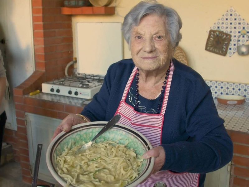 Eine der Pasta Grannies zeigt stolz ihre selbstgemachten Nudeln, die sie in einer großen Schüssel in den Händen hält.