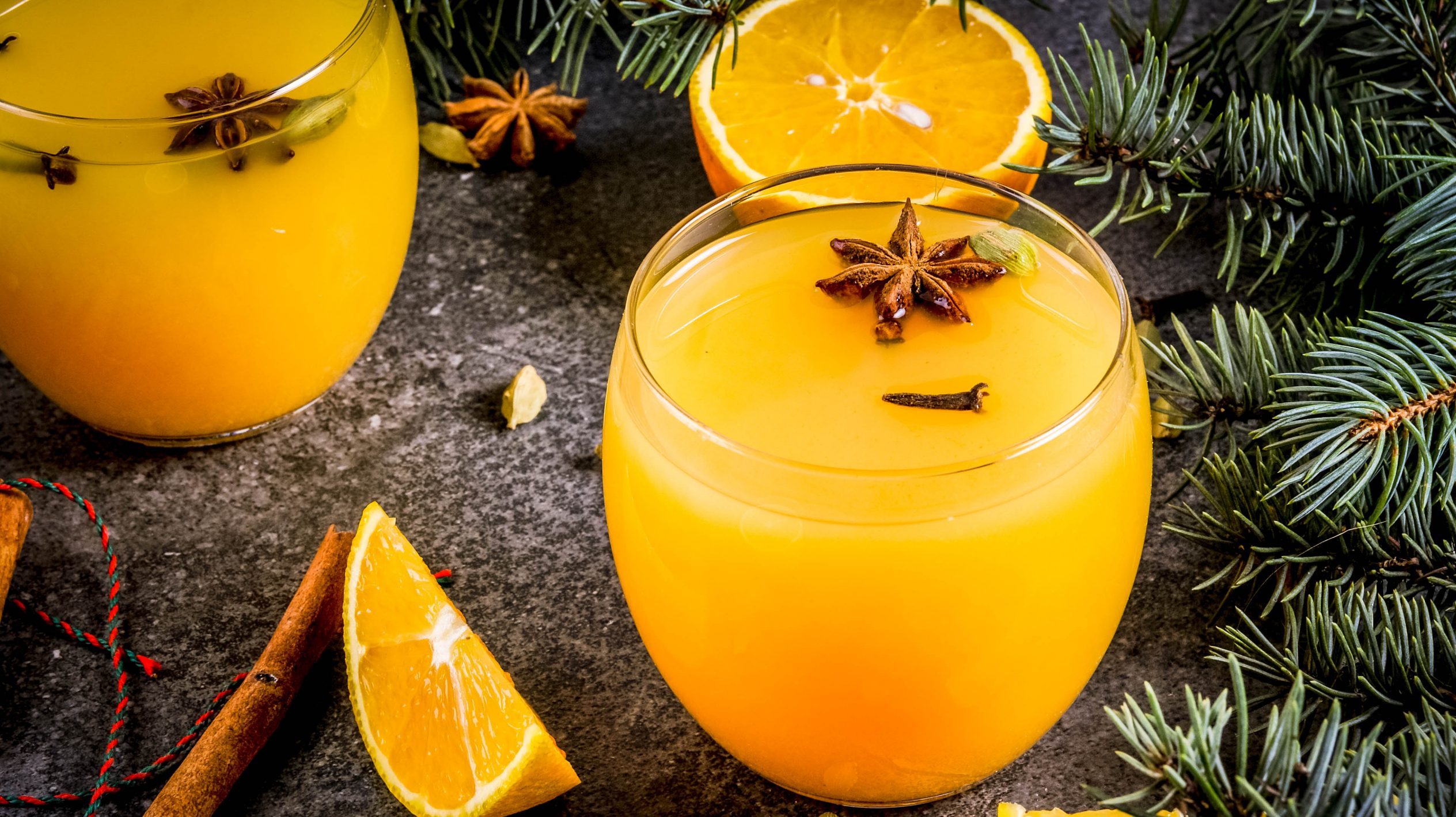 Der Weihnachts-Cocktail mit Orange und Zimt in zwei runden Gläsern angerichtet. Dazu sind im Hintergrund Teile eines Nadelbaums zu sehen.