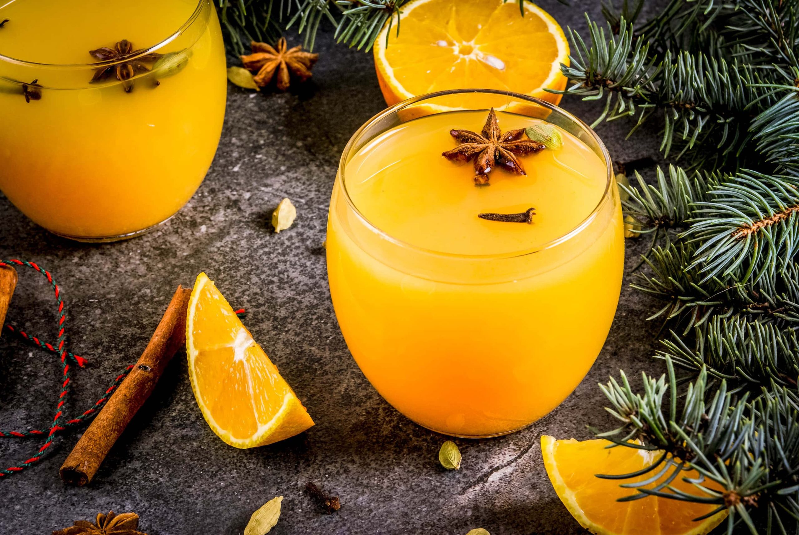 Der Weihnachts-Cocktail mit Orange und Zimt in zwei runden Gläsern angerichtet. Dazu sind im Hintergrund Teile eines Nadelbaums zu sehen.