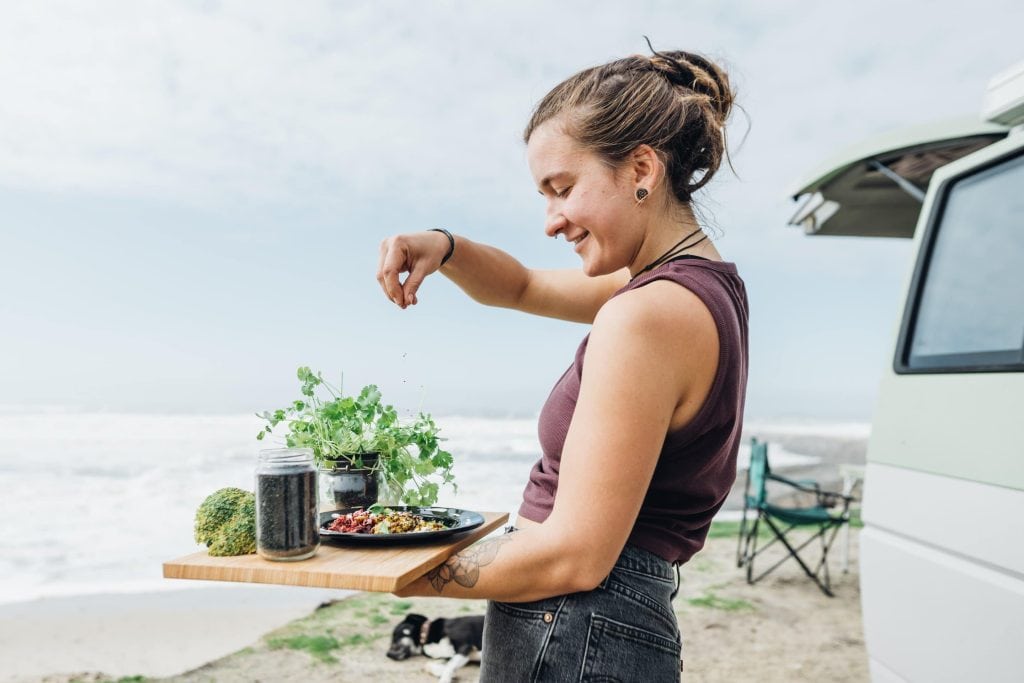 Die Autorin von Vegan im Van, Lorena, steht mit einem Brett vor ihrem Van, darauf steht ein gekochtes Essen mit frischen Kräutern. Im Hintergrund sieht man das Meer.