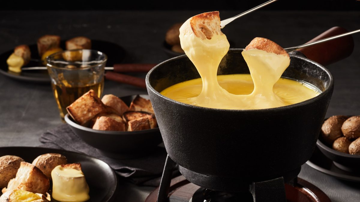Auf einem Tisch steht ein großer Topf mit Käse-Fondue. Zwei Spieße mit Brot und Käse sind über dem Topf.