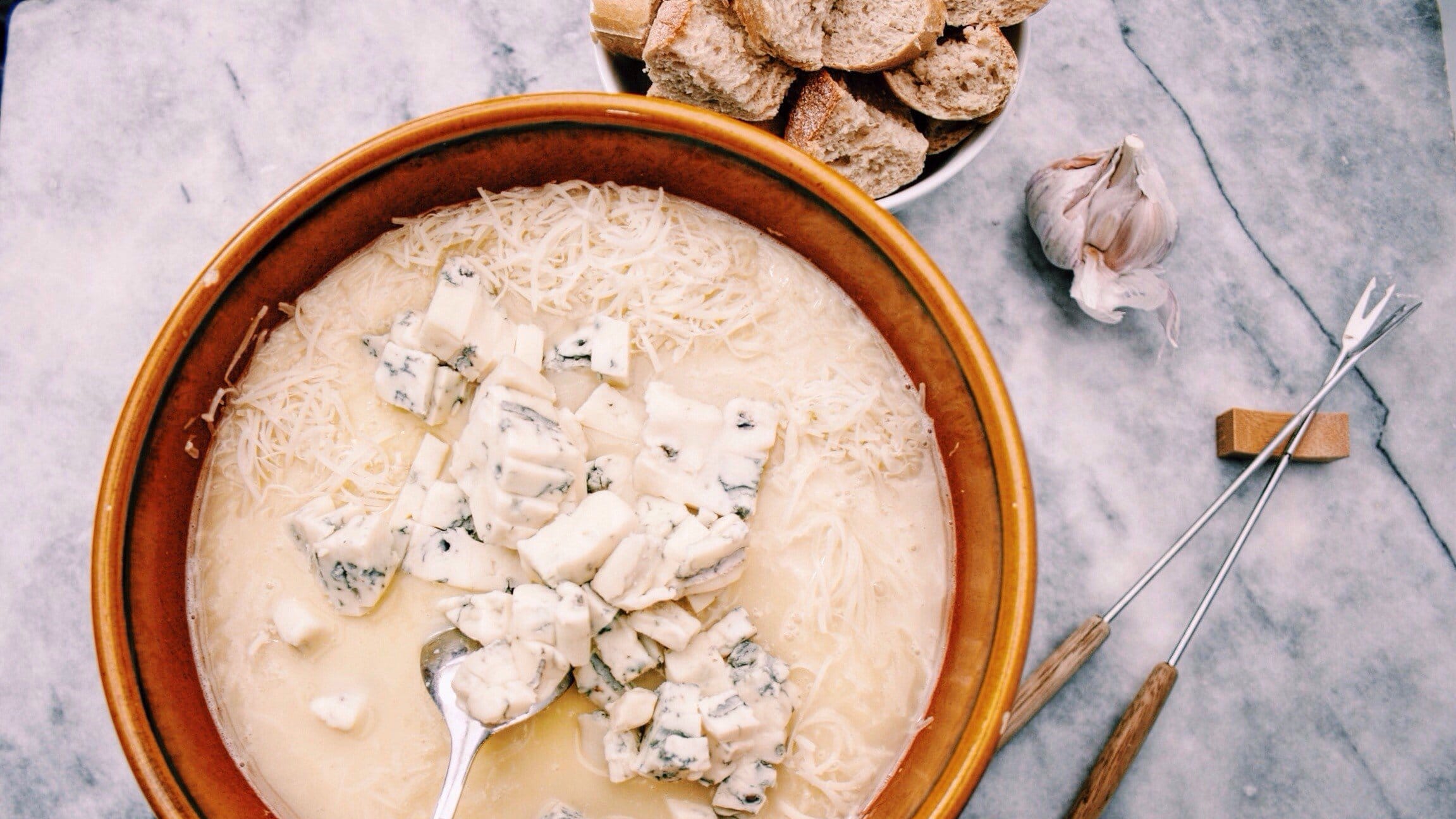 Käsefondue mit Gorgonzola im Tontopf auf Marmoruntergrund. Umgeben ist das Ganz von Brot, Knoblauch und Spießen.