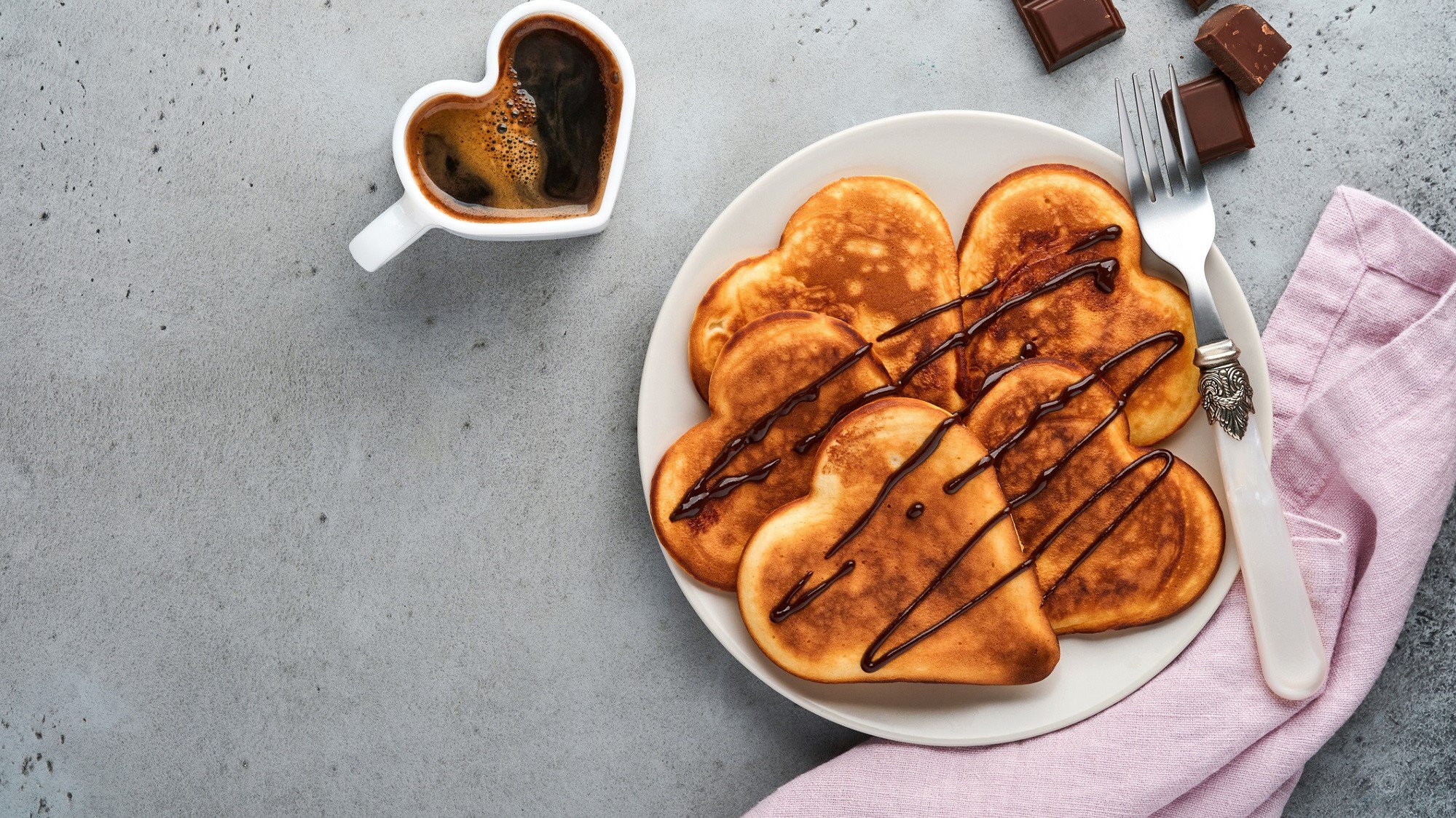 Auf einem Teller liegen herzförmige Buttermilch-Pfannkuchen mit Schokolasdensauce. Daneben steht eine Tasse Kaffee.