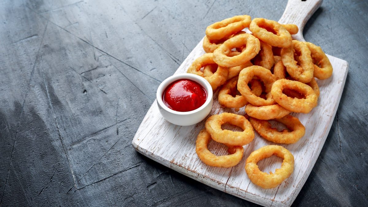 Onion Rings mit Ketchup auf Holzbrett, angerichtet auf grauem Untergrund