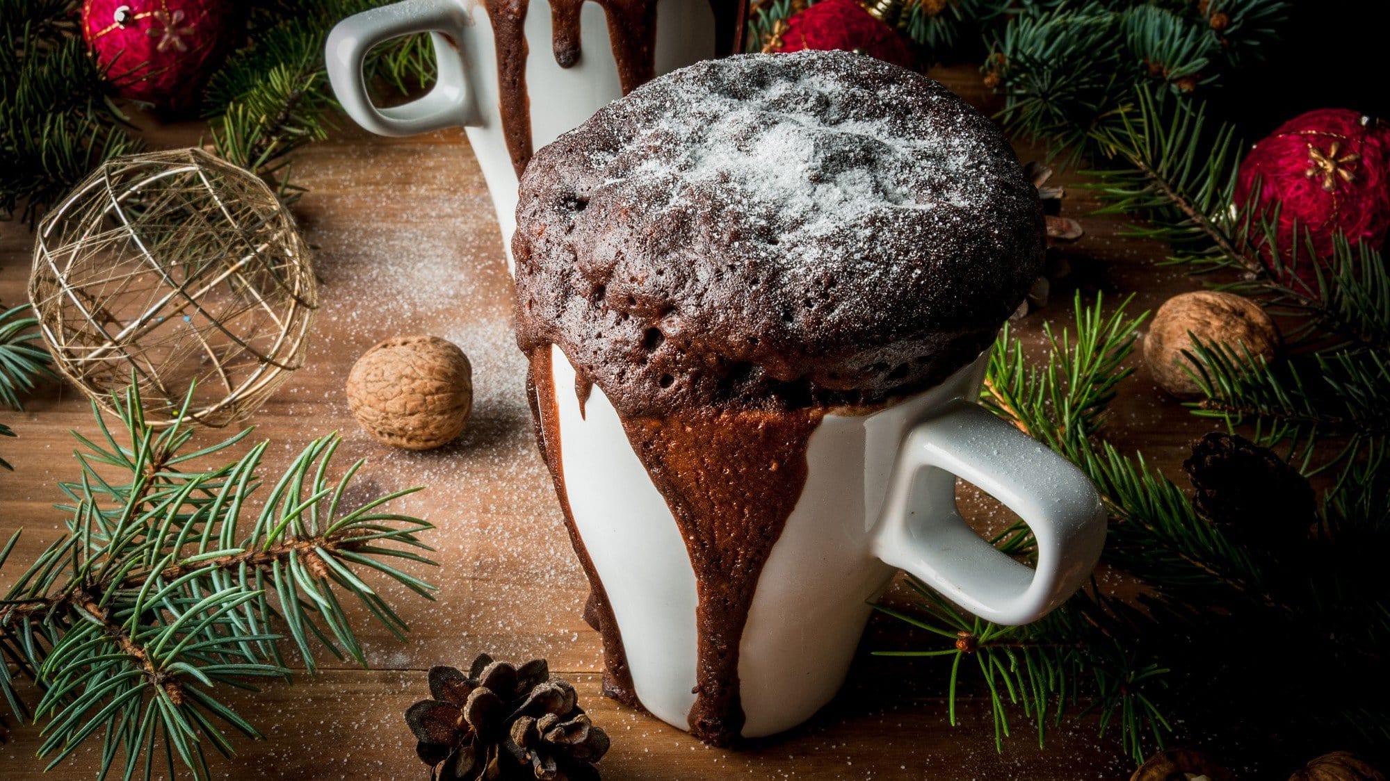 In einer weißen Tasse ist ein schokoladiger Tassenkuchen aufgebacken. Er ist mit Puderzucker bestreut. Die Tasse steht auf einem Holztisch, welcher weihnachtlich dekoriert ist.