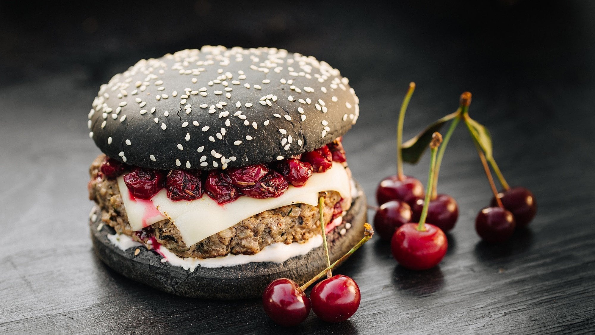 Ein Burger mit Sauerkirschen steht auf einem dunklen Holztisch. Der Burger besteht aus schwarzen Burgerbuns, Rindfleischpatties, Käse und Sauerkirschen.