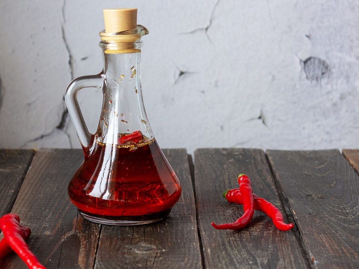 In einer durchsichtigen Karaffe aus Glas ist selbstgemachtes Chili-Öl. Drum herum liegen rote Chili-Schoten.