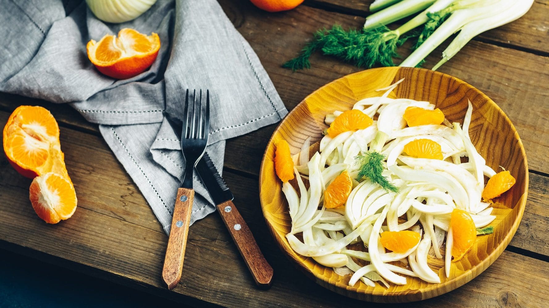Auf einem Holzteller ist eine Fenchel-Orangen-Salat mit Roquefort-Vinaigrette angerichtet. Neben dem Teller liegen Orangenstücke, ein Messer sowie eine Fenchelknolle.
