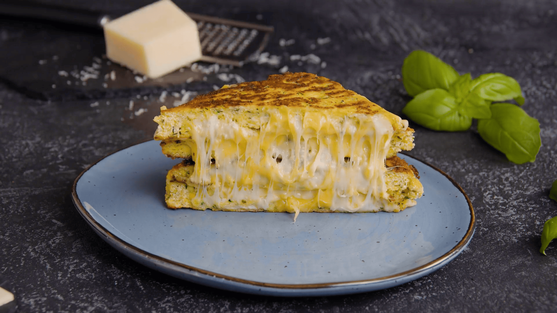 Auf einem blauen Teller liegt halbiertes ein Grilled Cheese mit Zucchinipuffern, dahinter einige Basilikumblätter und Parmesan.