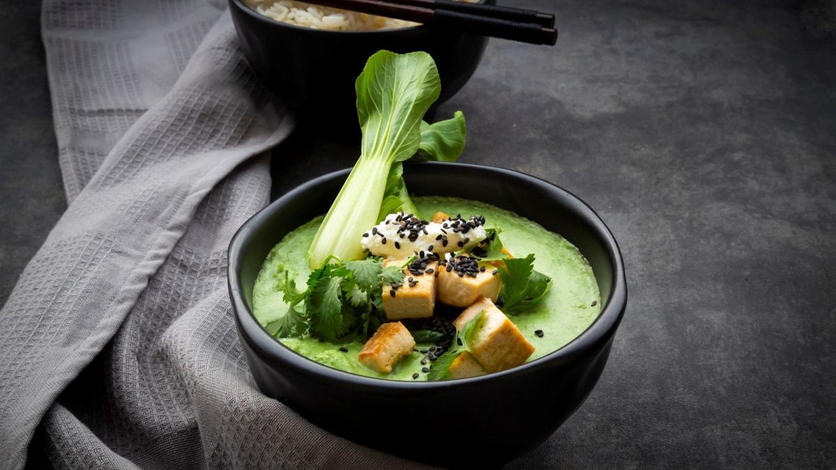 Eine schwarze Schüssel grünes Thai-Curry mit Pak Choi und Tofu steht auf einem dunkelgrauen Untergrund. Im Hintergrund steht eine Schüssel mit Reis und zwei Essstäbchen. Links neben dem Curry liegt ein graues Geschirrtuch.