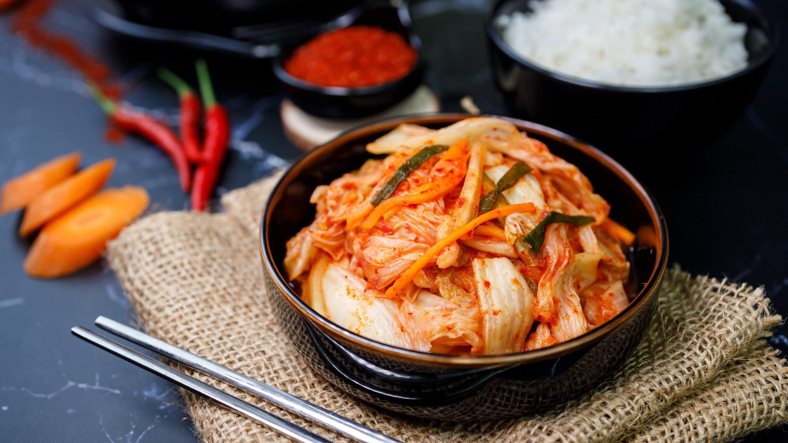 Eingelegter koreanischer Rettich und Kohl, auch Kimchi genannt, auf Reis in einer schwarzen Tonschale, gelegt auf ein Bastnetz mit Reis, Chilli, Karotte und Paprikapulver im Hintergrund.