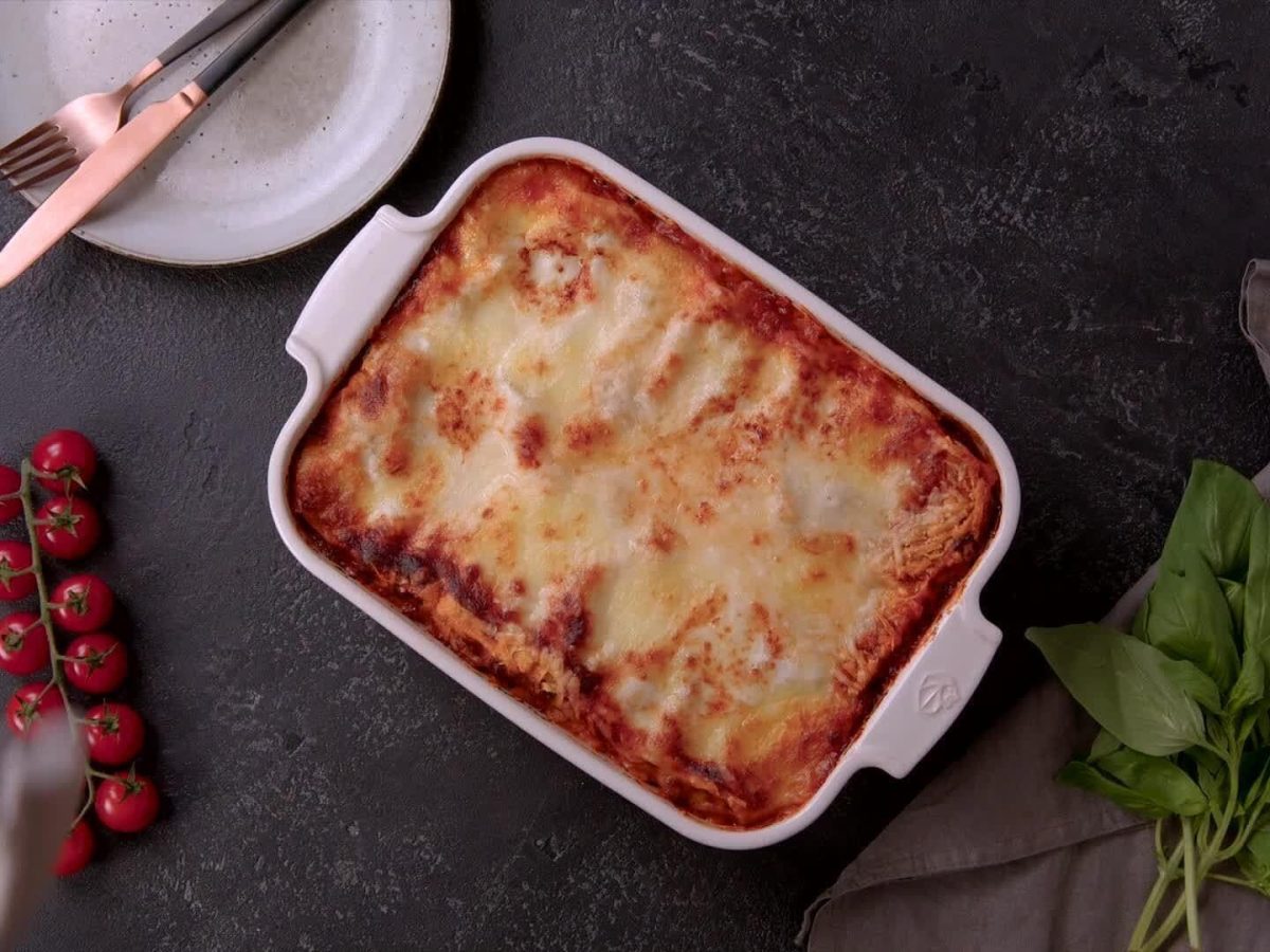 Unsere Lasagne frisch aus dem Ofen steht in einer weißen Auflaufform auf schwarzem Untergrund. Dazu ein Teller mit Besteck, ein Tomatenstrauch und Basilikum.
