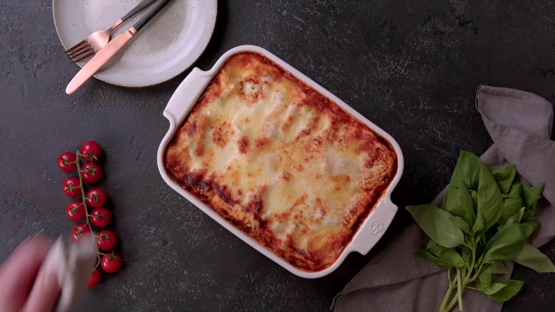 Unsere Lasagne frisch aus dem Ofen steht in einer weißen Auflaufform auf schwarzem Untergrund. Dazu ein Teller mit Besteck, ein Tomatenstrauch und Basilikum.