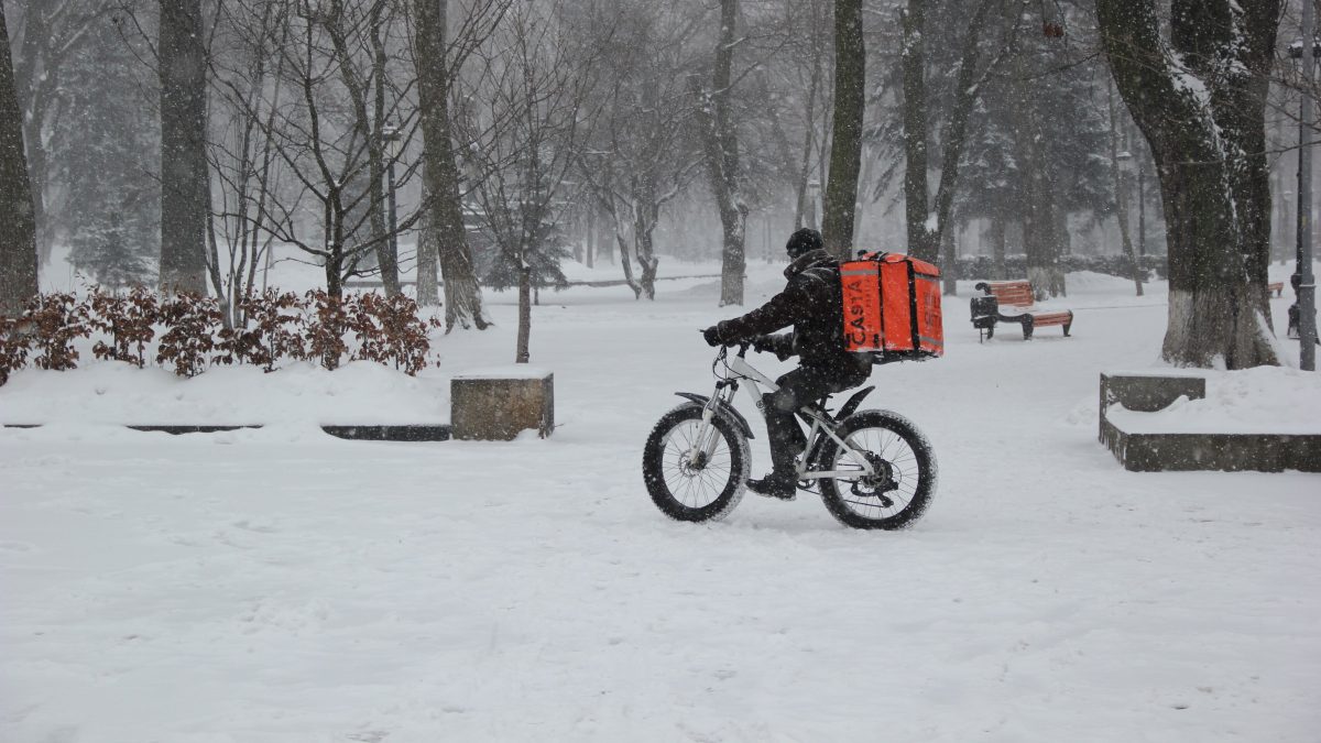 Lieferant mit Rucksack auf Fahhrad fährt durch einen schneebedeckten Park bei Schneefall. Aufnahme aus der Ferne.