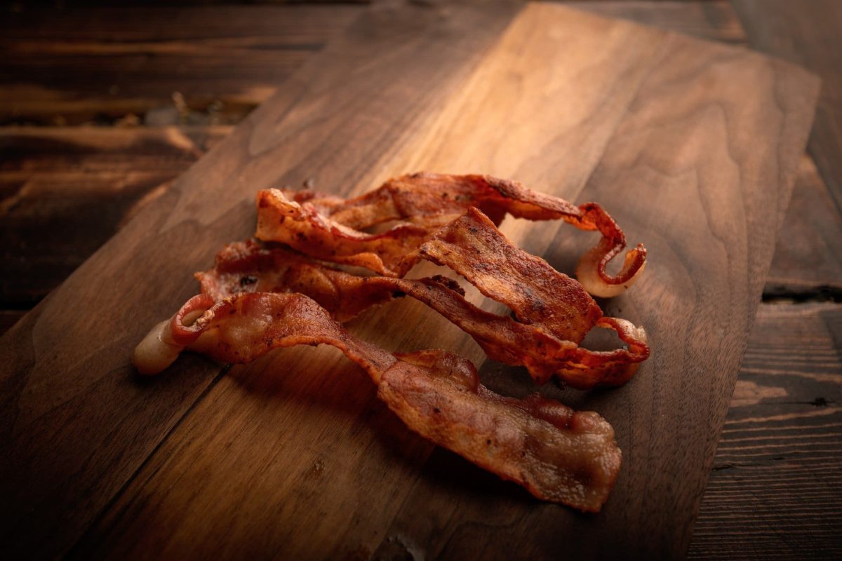 Zum Tag des Bacons: Vier Scheiben Bacon auf Holzbrett auf Holztisch mit dunkelbrauner Maserung. Aufnahme aus der Draufsicht.