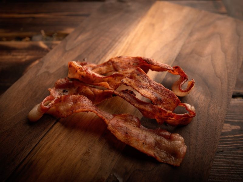 Zum Tag des Bacons: Vier Scheiben Bacon auf Holzbrett auf Holztisch mit dunkelbrauner Maserung. Aufnahme aus der Draufsicht.