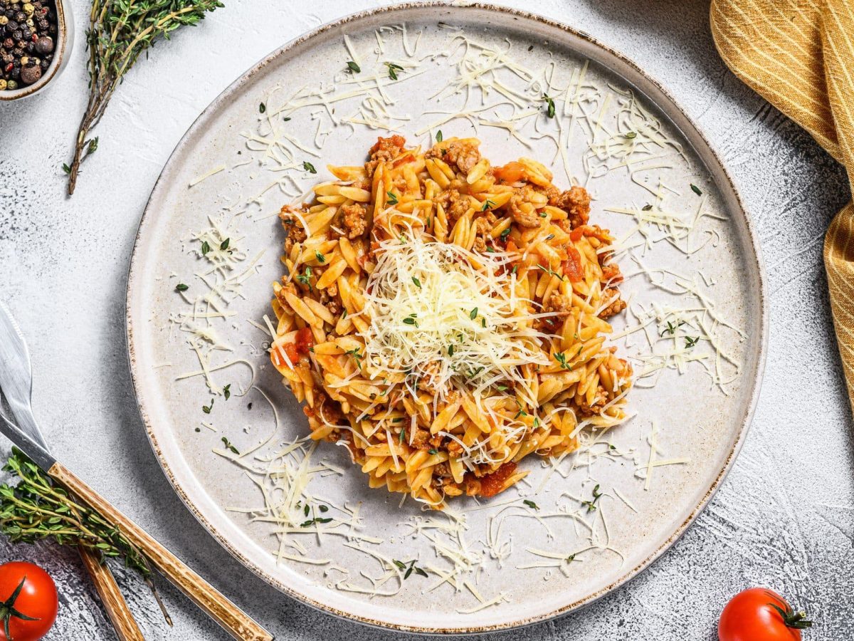 Ein Teller veganes Orzorisotto in der Draufsicht, mit veganem Parmesan bestreut, daneben Besteckt, ein Geschirrtuch, Tomaten und Gewürze.