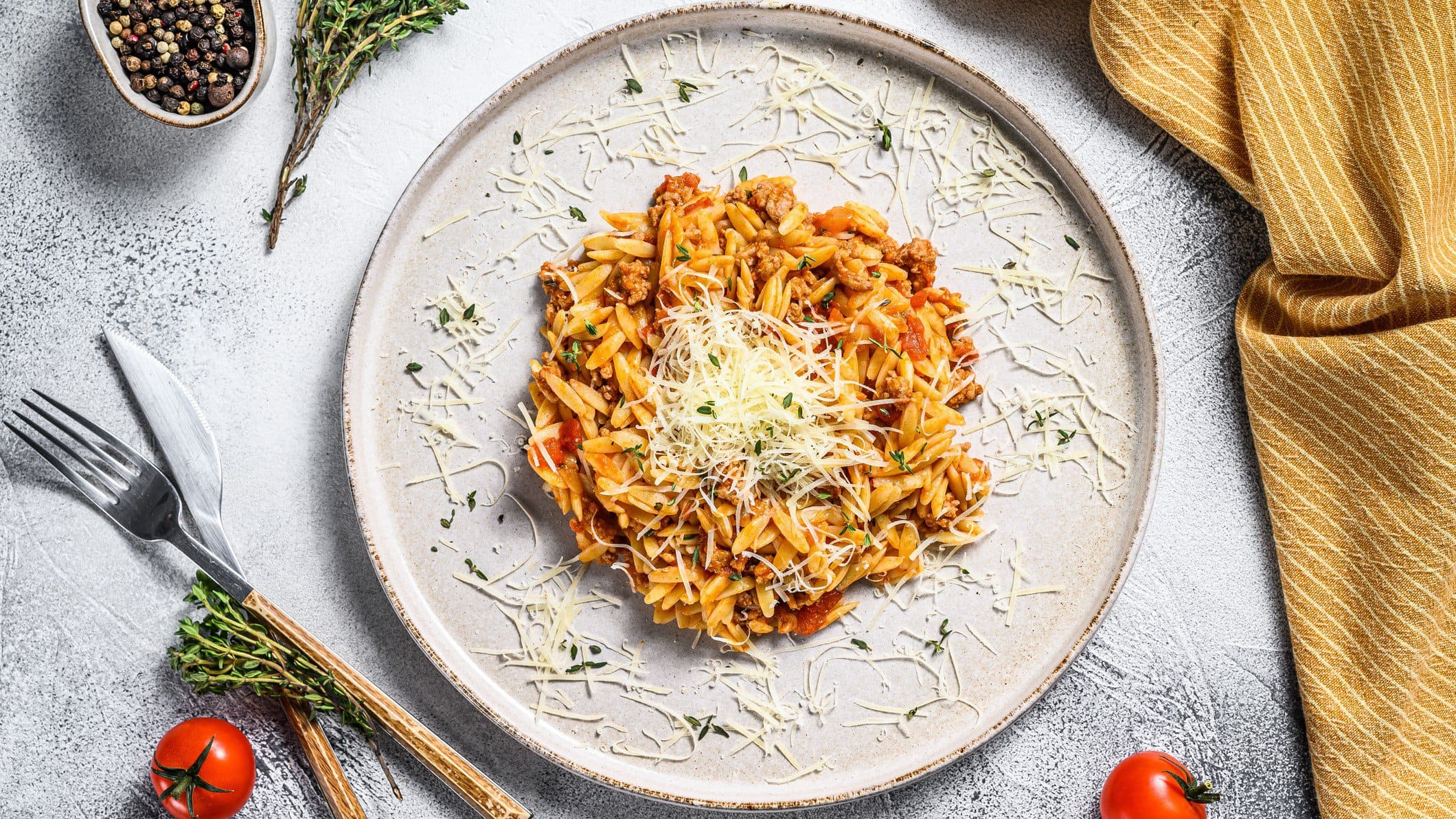 Ein Teller veganes Orzorisotto in der Draufsicht, mit veganem Parmesan bestreut, daneben Besteckt, ein Geschirrtuch, Tomaten und Gewürze.