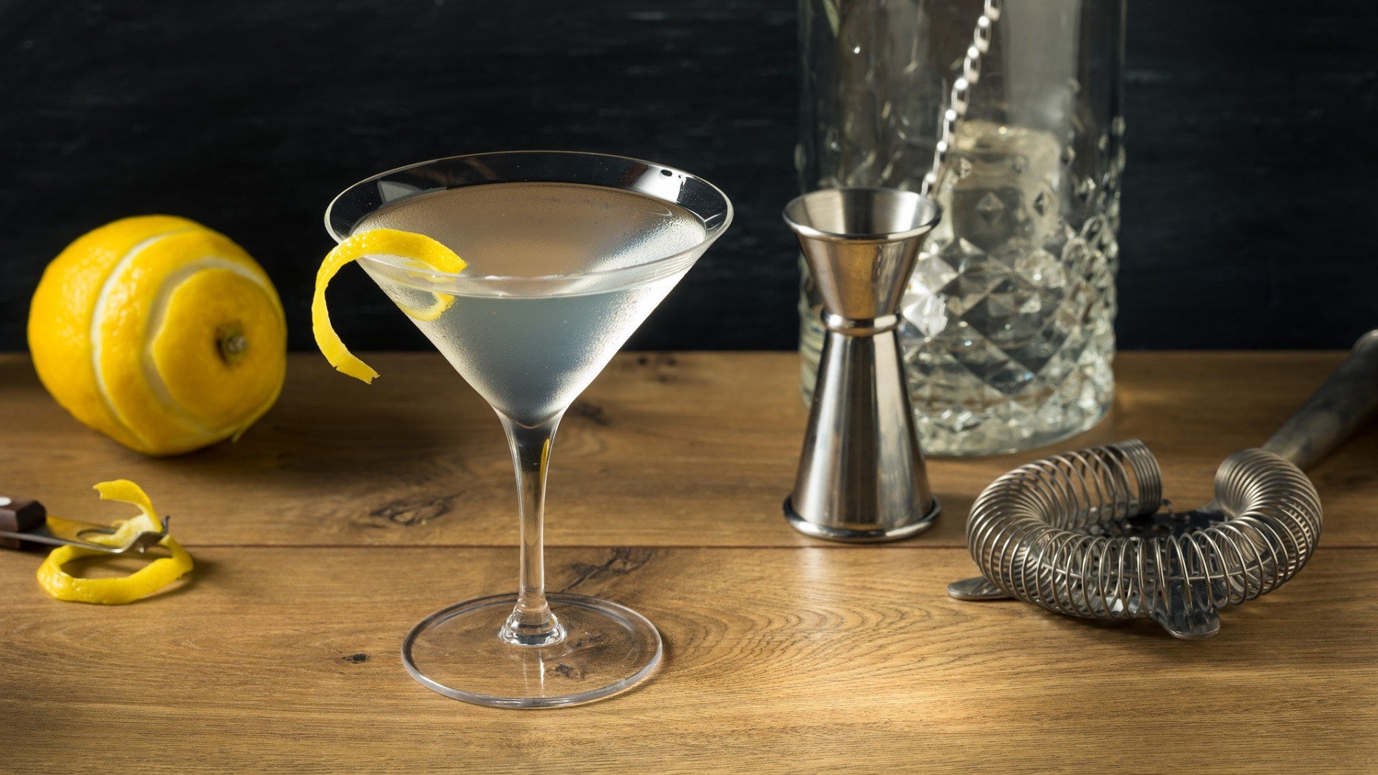Ein Vesper Cocktail in einem Martini Glas steht auf einem dunklen Holztisch. Im Hintergrund liegen verschiedene Utensilien zum Mixen von Cocktails und eine Zitrone.