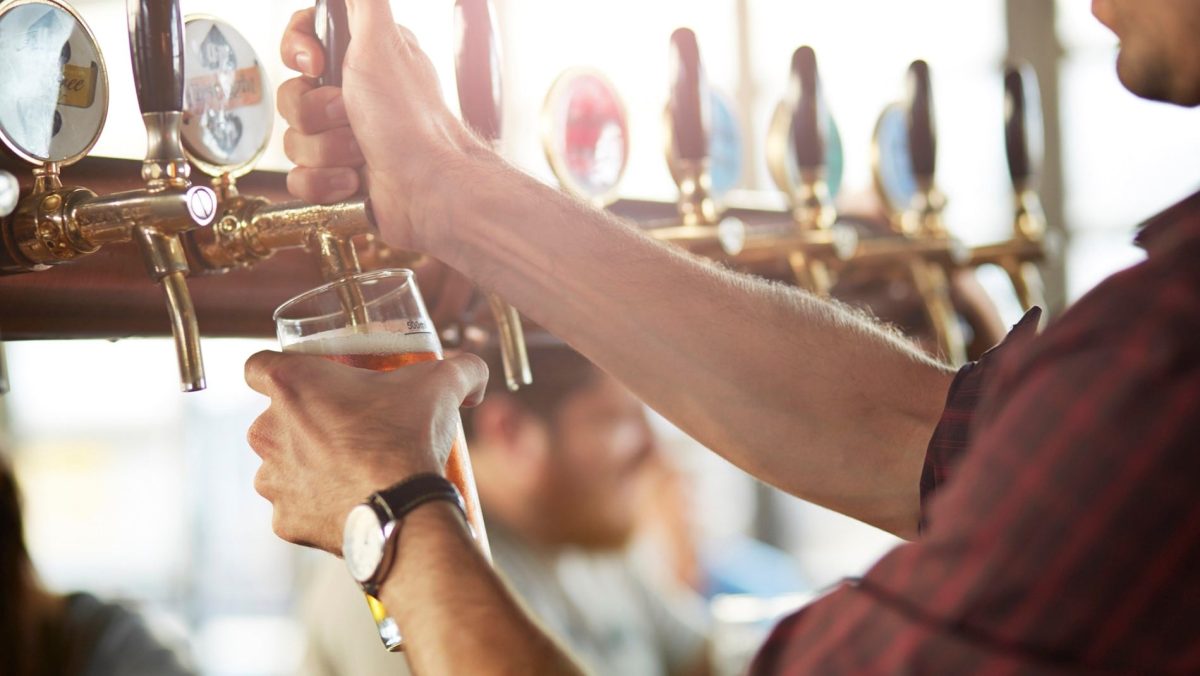Ein Mann in einem karierten Hemd zapft helles Bier in ein großes Glas. Im Hintergrund sitzen mehrere Personen an einer Bar.