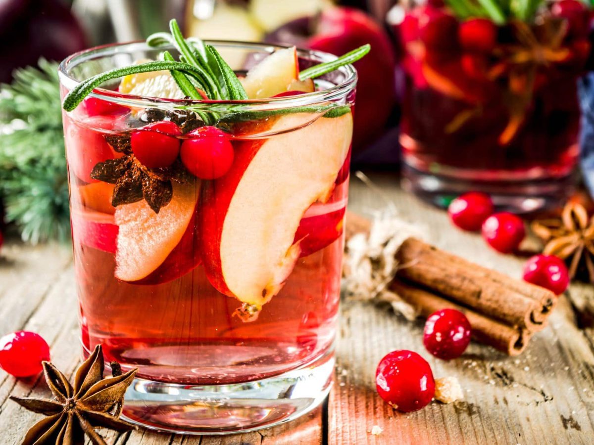 Ein Glas winterliche Sangria mit Apfel und Orange sowie Cranberrys, Anis und Rosmarin. Im Hintergrund sind unscharf die Zutaten zu erkennen.