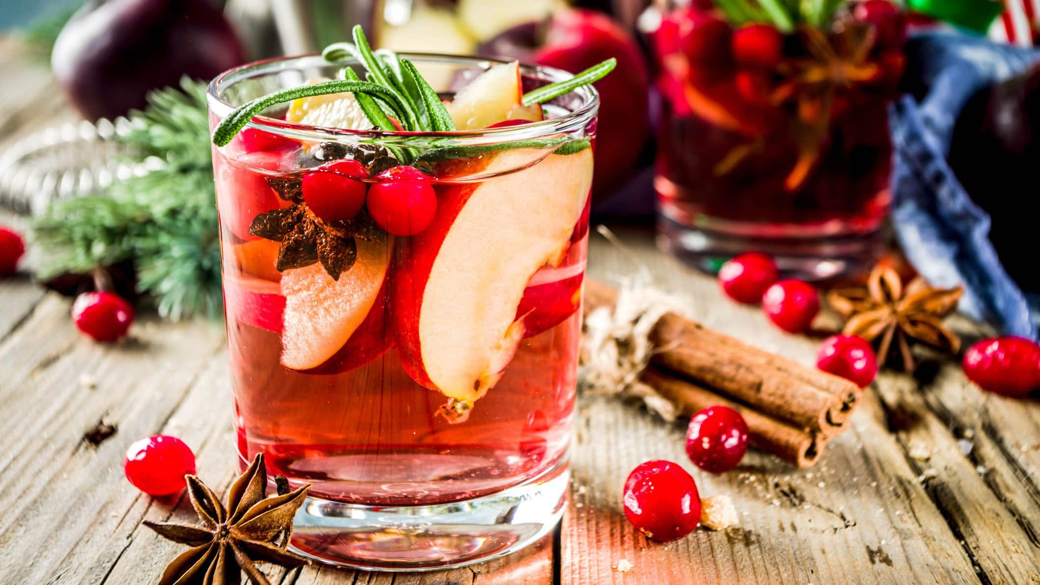Ein Glas winterliche Sangria mit Apfel und Orange sowie Cranberrys, Anis und Rosmarin. Im Hintergrund sind unscharf die Zutaten zu erkennen.