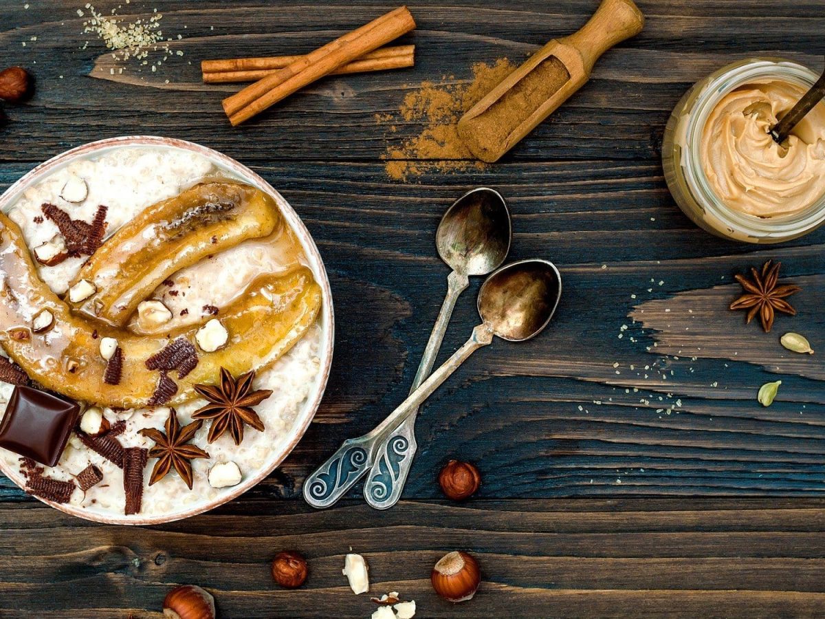 Ein winterlicher Porridge mit gebackenen Bananen steht auf einem dunklen Untergrund. Der Porridge ist angerichtet mit Schokolade, Zimt und Sternanis. Auf dem Tisch verteilt liegen Nüssem Löffel und Gewürze.