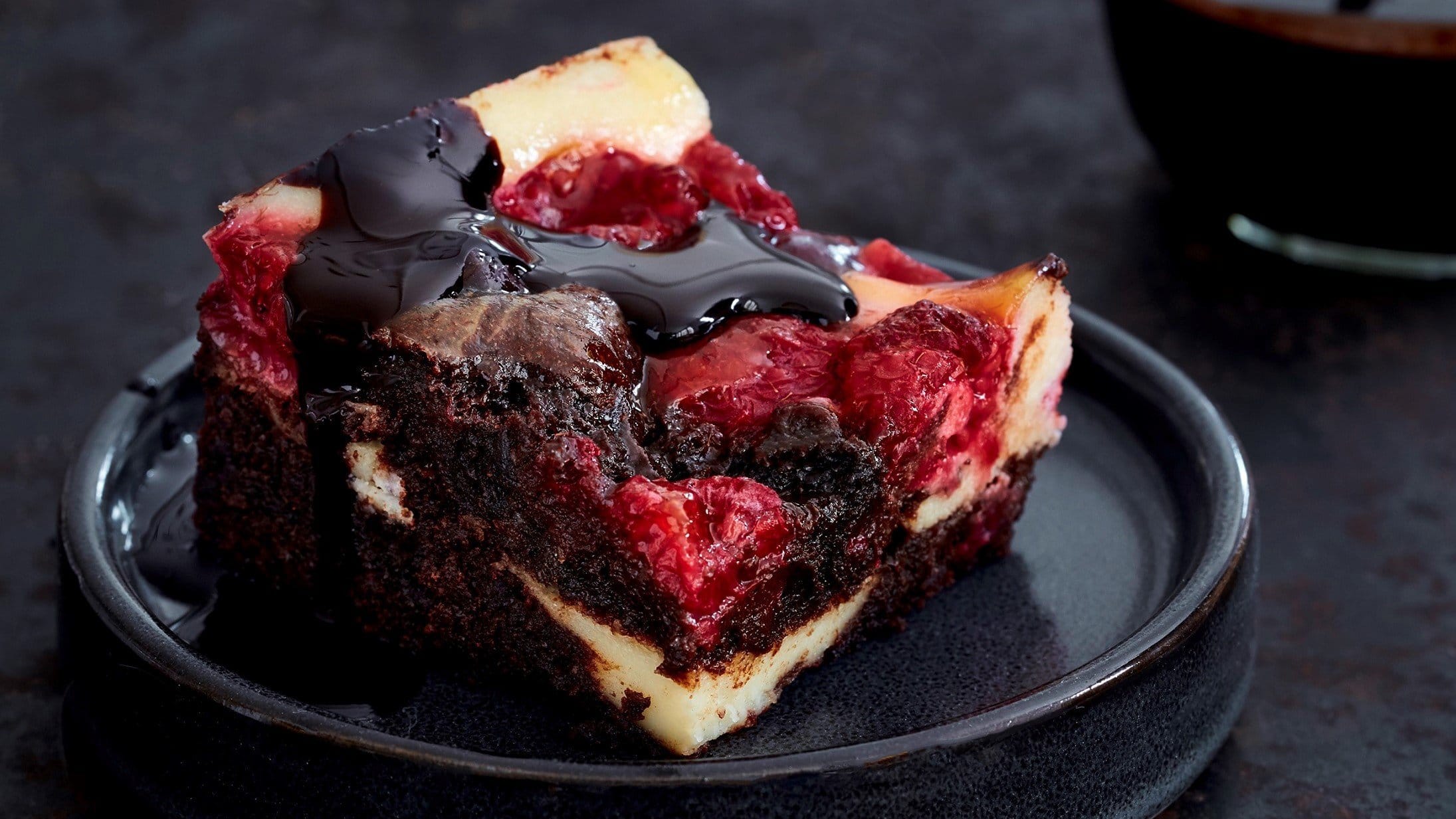 Auf einem schwarzen Porzellan-Teller liegt ein Stück der Beeren-Cheesecake-Brownies. Es ist überzogen mit Schokoladensoße.