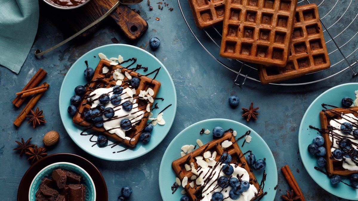 Auf drei blauen Tellern sind schokoladige Brownie Waffeln verteilt. Sie sind dekoriert mit Blaubeeren, Naturjoghurt und Schokoladensauce.