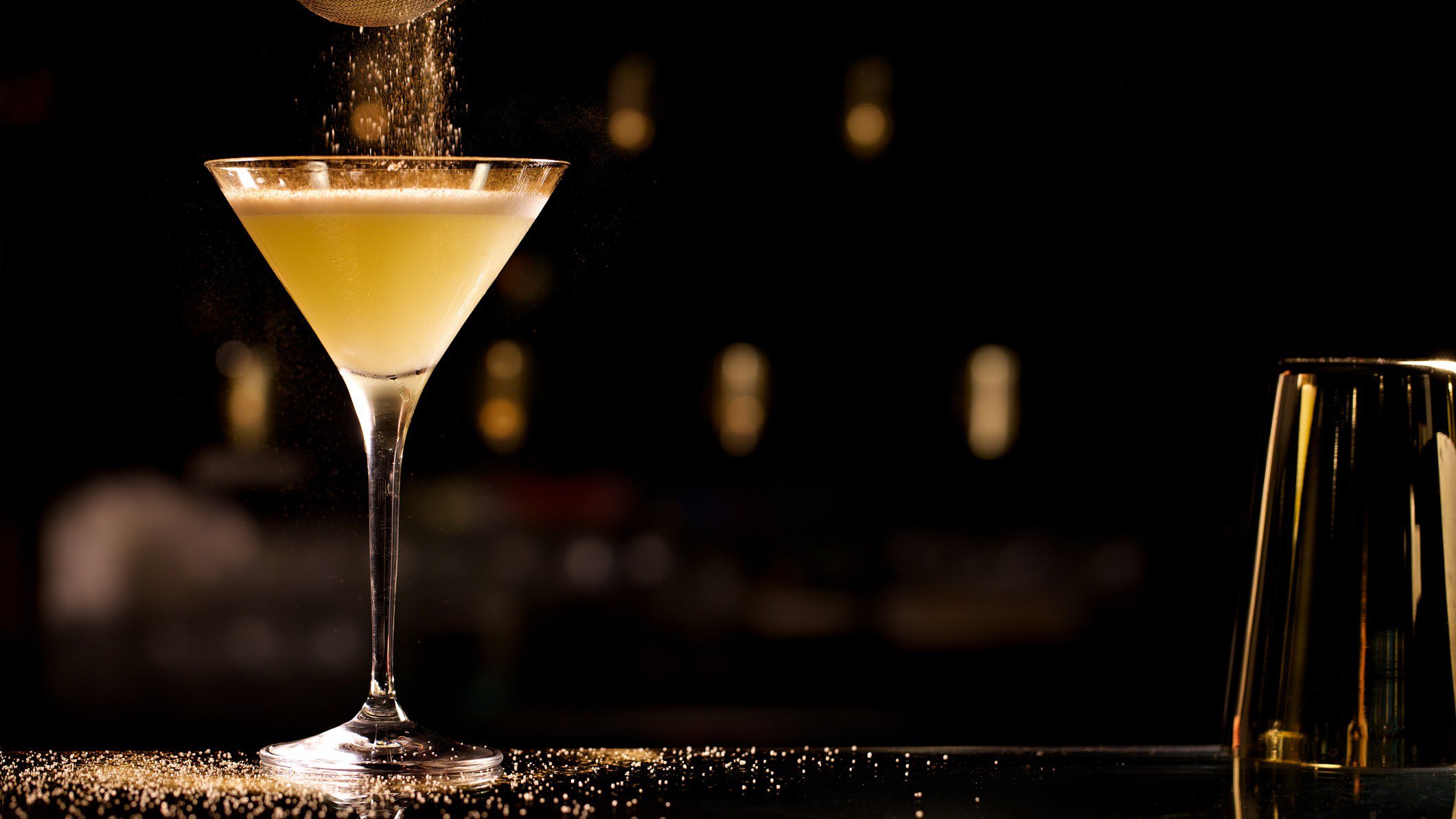 French 75 aus "Casablanca" Champagner-Cocktail in Champagnerglas auf dunklem Tisch vor dunklem Hintergrund. Zucker rieselt von oben in das Glas. Ein weiteres Glas im Vordergrund angedeutet. Frontalaufnahme.