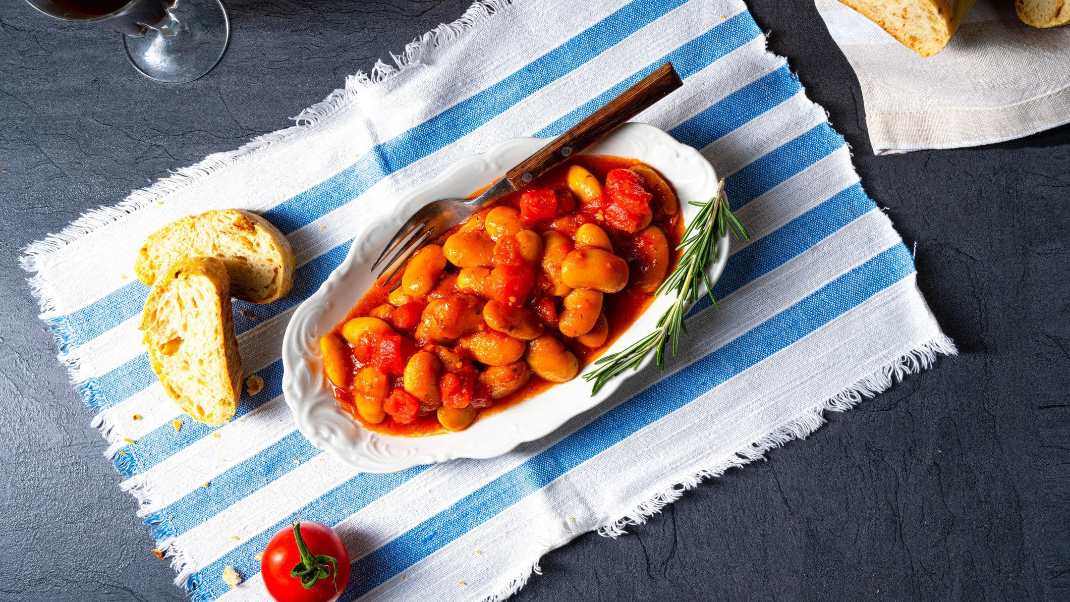 Weißer Teller mit griechischen Bohnen in Tomatensauce "Gigantes Plaki" auf gestreiftem Tischtuch.