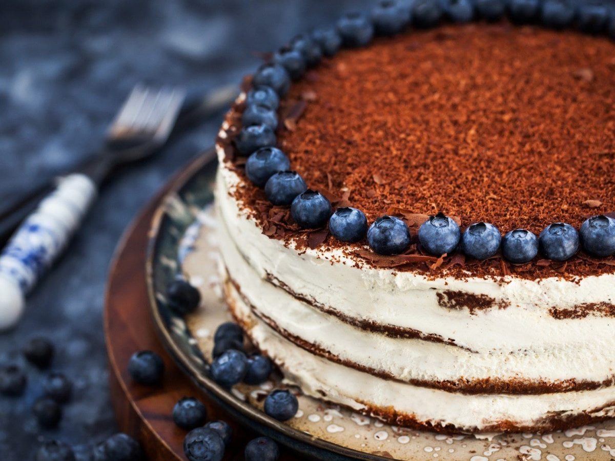 Auf einem Teller steht ein Naked Cake mit Blaubeeren am Rande des Kuchens. Er ist mit Schokoraspeln bestreut. Daneben liegt Besteck.