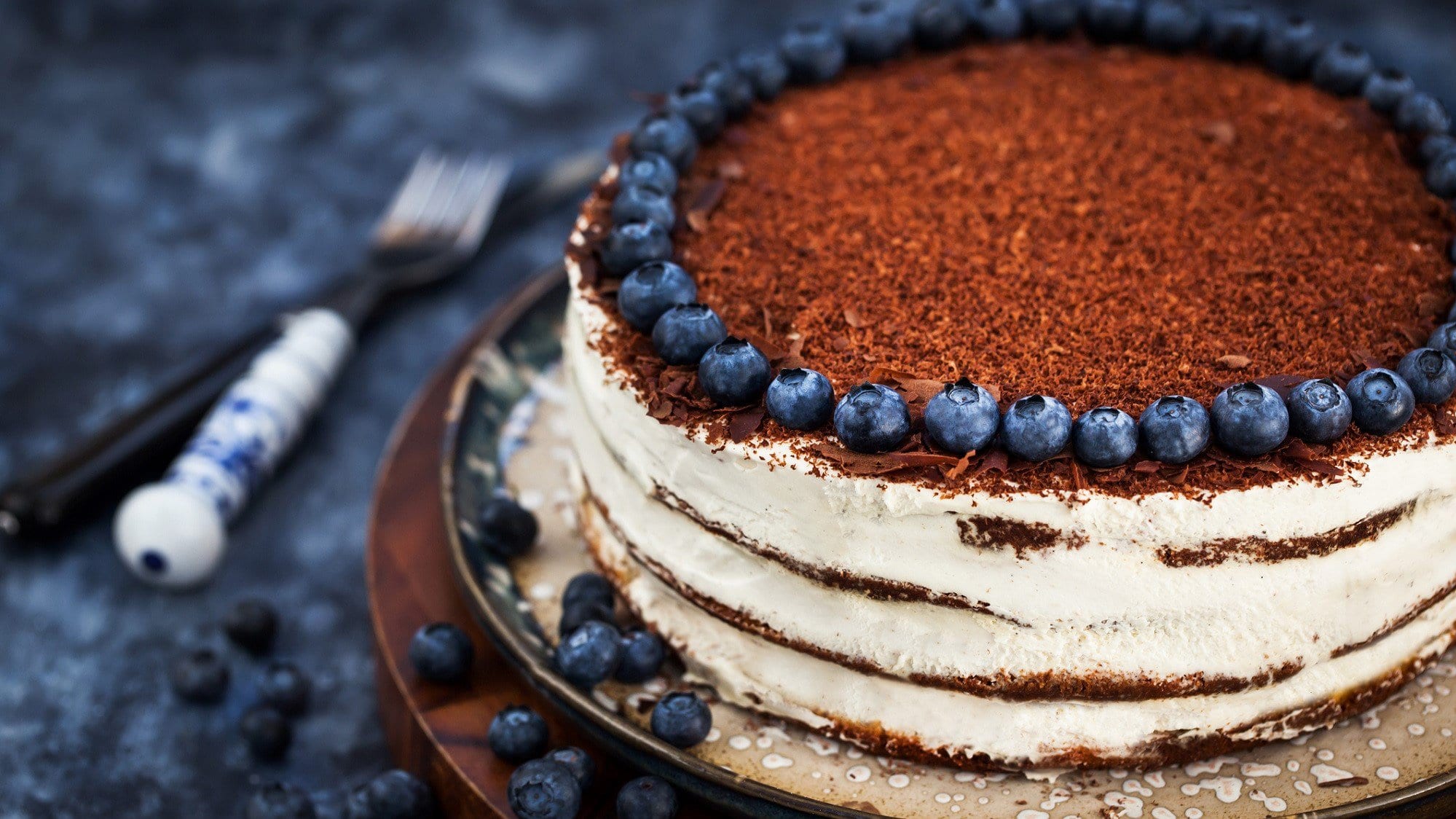 Auf einem Teller steht ein Naked Cake mit Blaubeeren am Rande des Kuchens. Er ist mit Schokoraspeln bestreut. Daneben liegt Besteck.