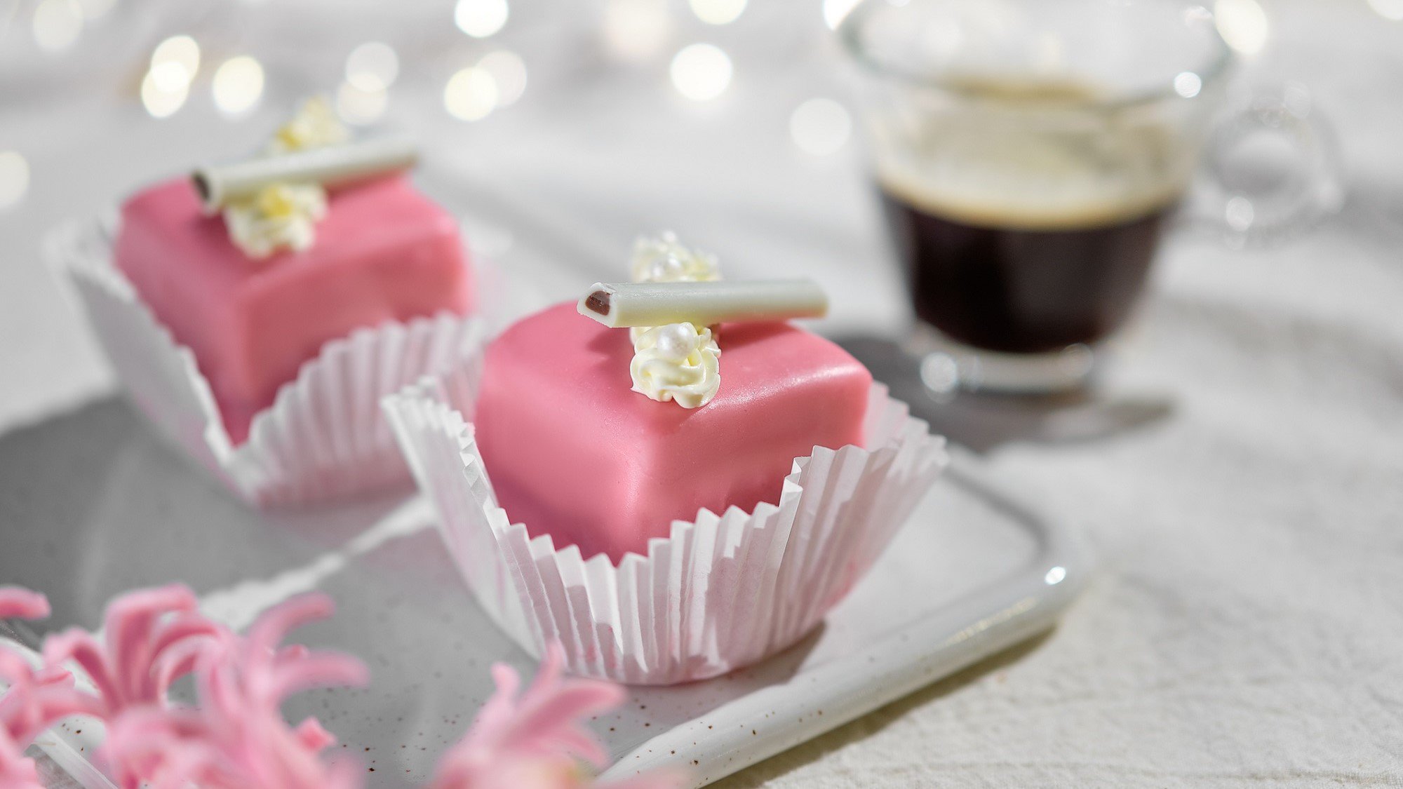 In einer kleinen Papierform sind Petit Fours angerichtet. Sie sind mit rosa Marzipan überzogen und mit Zuckerperlen dekoriert.
