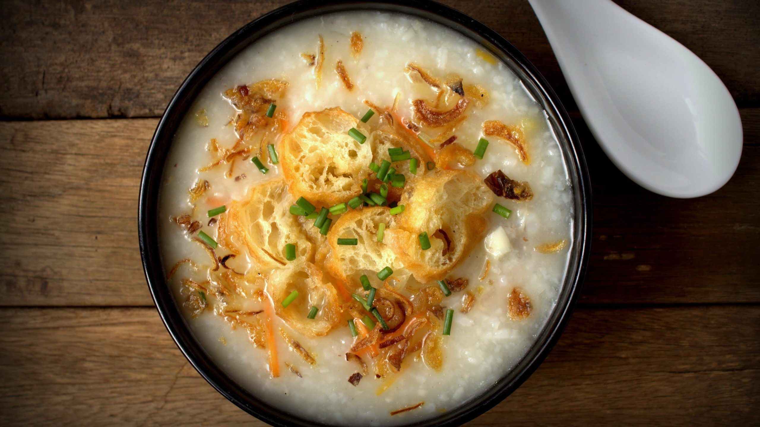 Asiatische Reissuppe, auch Congee genannt, mit Reis, Gewürzen, garniert mit frittierten Teigstangen, Schnittlauch und Röstzwiebeln in einer schwarzen Schale auf hölzernem Untergrund
