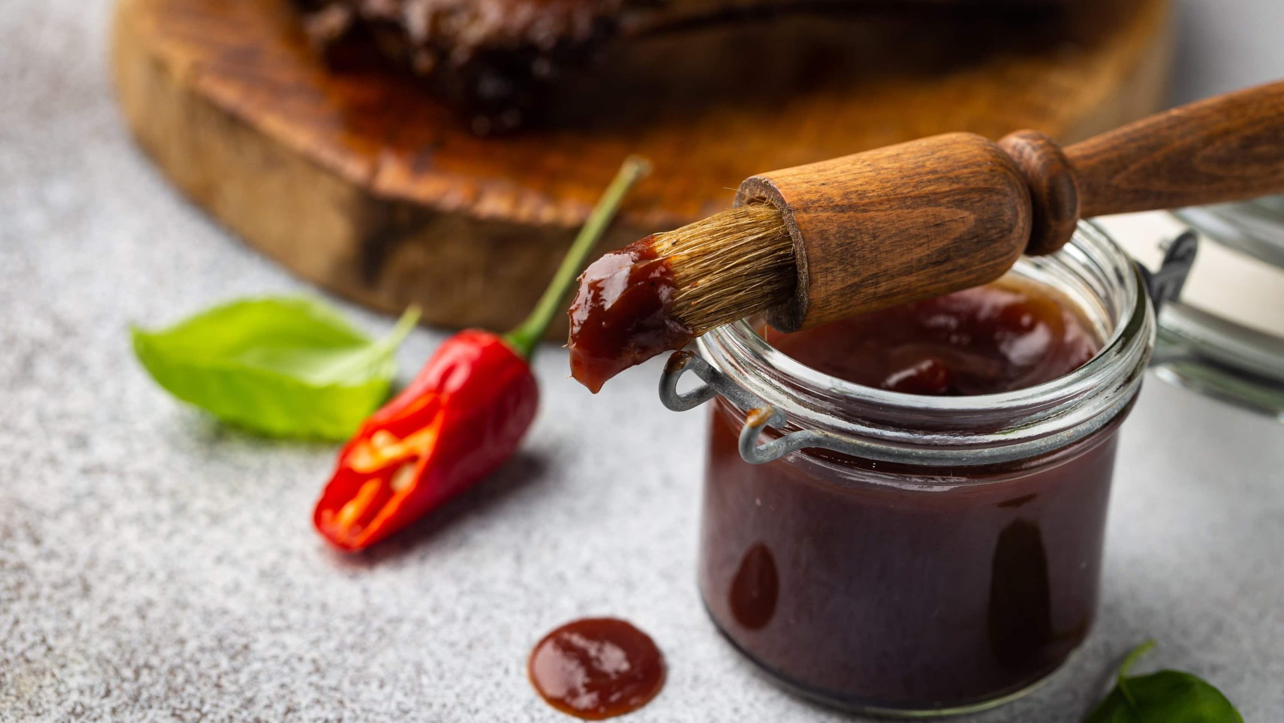 Basting Marinade und Sauce im Glas mit Glasur-Pinsel. Chili und Fleisch auf einem Holzbrett im Hintergrund. Frontalansicht.