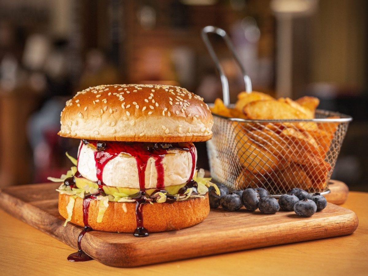Ein Burger mit Camembert und Blaubeersauce steht auf einem Holzbrett. Der Burger ist zusätzlich angerichtet mit Ananas und Salat. Um den Burger herum liegen Blaubeeren, am Rand des Bretts steht ein Sieb mit frittierten Kartoffeln.