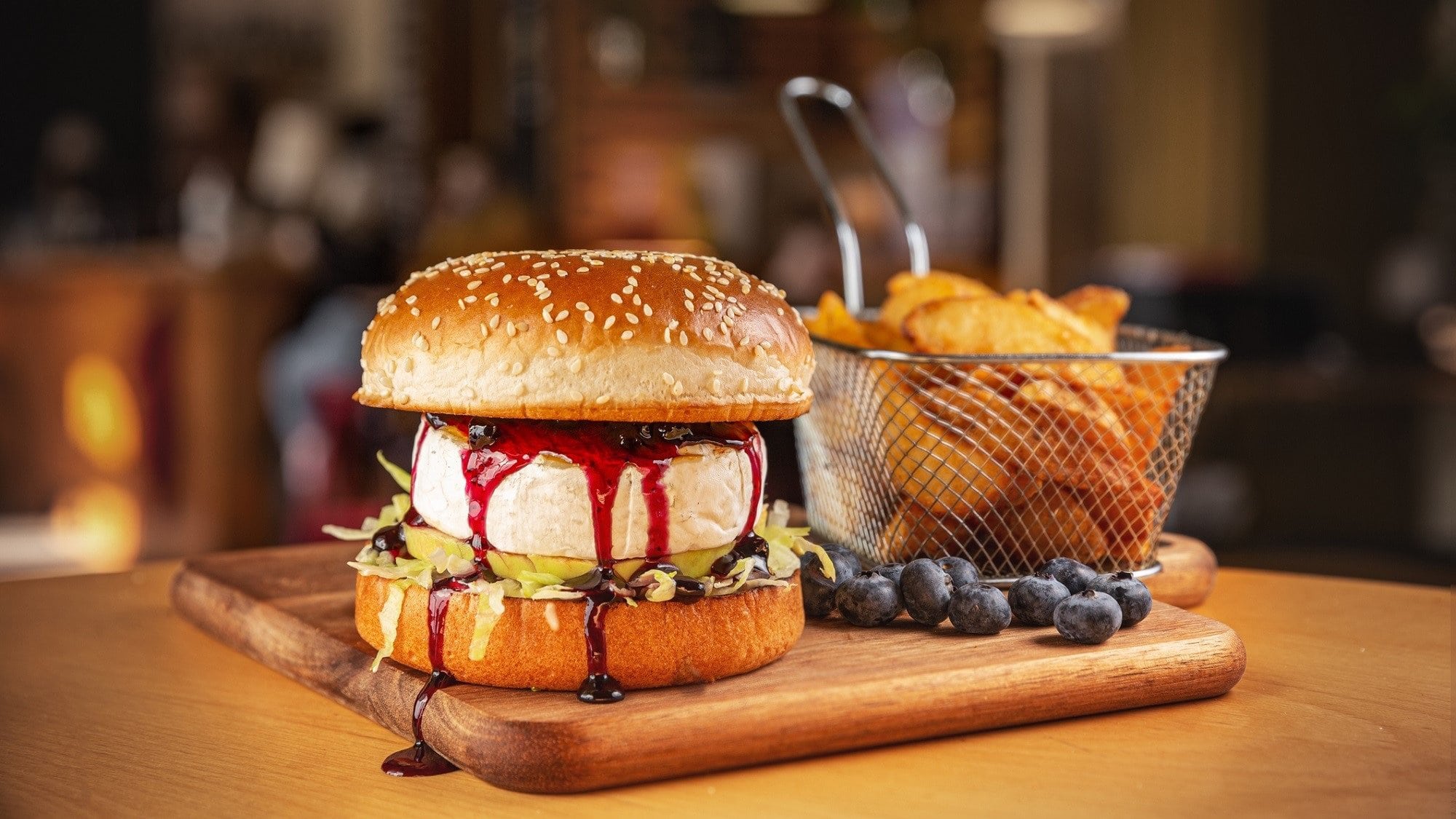 Ein Burger mit Camembert und Blaubeersauce steht auf einem Holzbrett. Der Burger ist zusätzlich angerichtet mit Ananas und Salat. Um den Burger herum liegen Blaubeeren, am Rand des Bretts steht ein Sieb mit frittierten Kartoffeln.