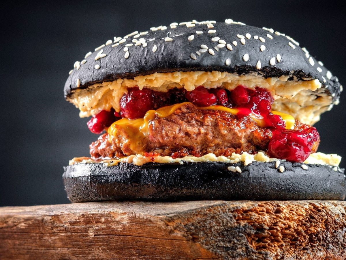 Ein Burger mit Cranberry-Sauce in schwarzen Burgerbuns steht auf einem dunklen Holzbrett vor einem schwarzen Hintergrund