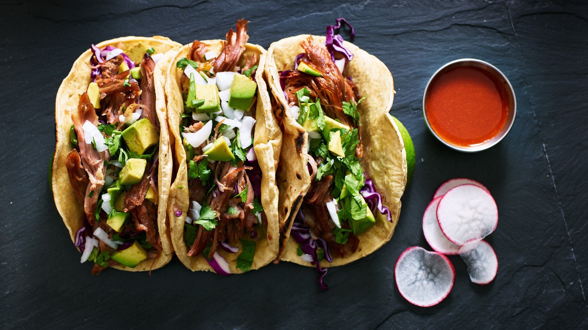 Dreimal Carnitas fuer Tacos Tacos mit Füllung auf schwarzem Tisch. Daneben Radieschen und eine Schüssel mit roter Sauce. Aufnahme aus der Vogelperspektive.