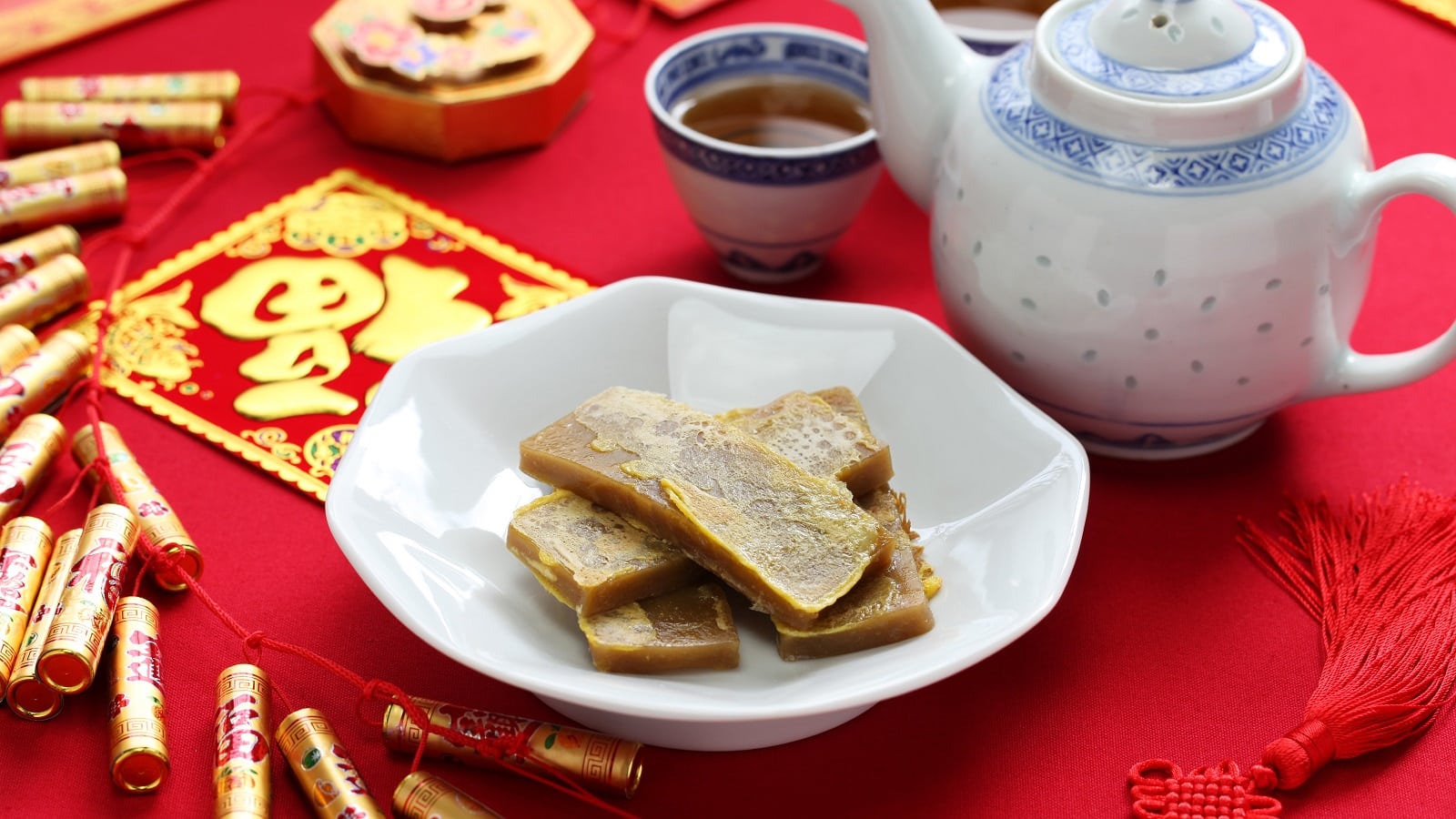 Eine weiße Porzellan-Schüssel mit mehreren Scheiben des chinesischen Reiskuchens Nian Gao neben einer Kanne und einer Tasse mit dunklem Tee, alles auf einem Tisch, der mit einem roten Tuch ausgelegt ist und mit festlichen Dekorationen verziert ist.